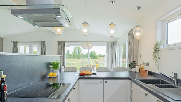 Een moderne keuken met een centraal eiland, afzuiging boven het kookoppervlak, hangende lampen en uitzicht op de omgeving.
