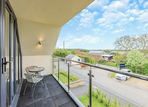 A balcony with a table and chairs, a view of a road with a car, and houses in the distance.