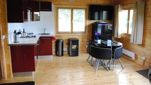 Una cocina compacta con paredes de madera, un comedor con mesa y sillas, un televisor montado sobre la encimera, y una ventana con persianas.