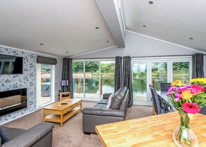 A modern living room with a sofa, a wooden table, a TV, and a view of a lake through large windows. - Gressingham 50 is a beautiful lodge in a fantastic location. (Carnforth)
