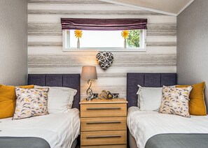 A bedroom with two beds, a dresser, a lamp, and a window with a view of greenery. - Gressingham 50 is a beautiful lodge in a fantastic location. (Carnforth)
