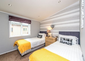 A hotel room with two beds, a window with blinds, a bedside table with a lamp, and a wall-mounted heart-shaped decoration. - Gressingham 50 is a beautiful lodge in a fantastic location. (Carnforth)