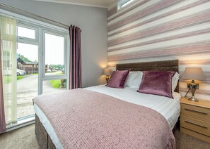 A bedroom with a large bed, a nightstand, a lamp, and a view of the outside. - Gressingham 50 is a beautiful lodge in a fantastic location. (Carnforth)