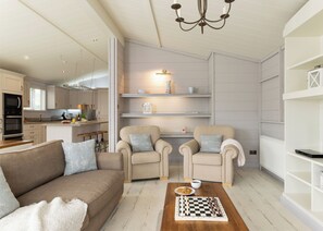A modern living room with a sofa, two armchairs, a coffee table with a chessboard, and a kitchen in the background. - 2 bedroom accommodation in Honiton (Honiton)