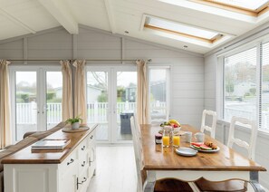 A dining area with a wooden table, chairs, and a view of the outdoors through large windows. - 2 bedroom accommodation in Honiton (Honiton)