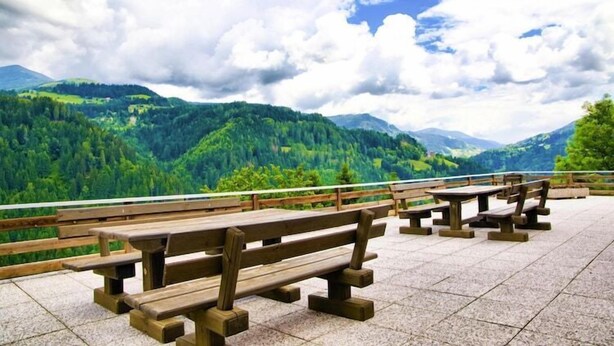 A scenic outdoor seating area with wooden benches and tables, overlooking a mountainous landscape.