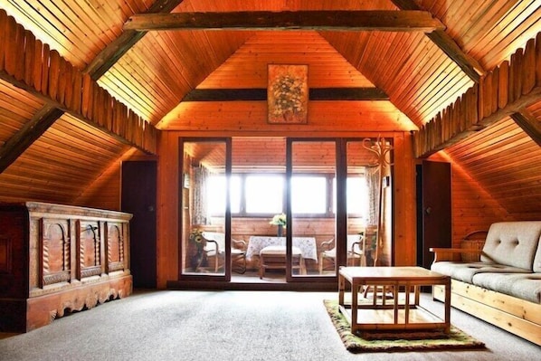 A cozy attic room with wooden beams, a large sliding glass door leading to a dining area, and a sofa.