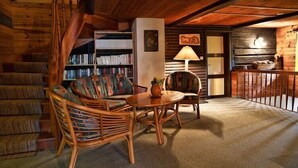 A cozy log cabin interior with a spiral staircase, a bookshelf, a dining area with wicker furniture, and a lamp.
