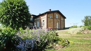 Ein großes, zweistöckiges Haus mit Backsteinfassade und zahlreichen Fenstern, umgeben von üppigem Grün und blühenden Pflanzen.