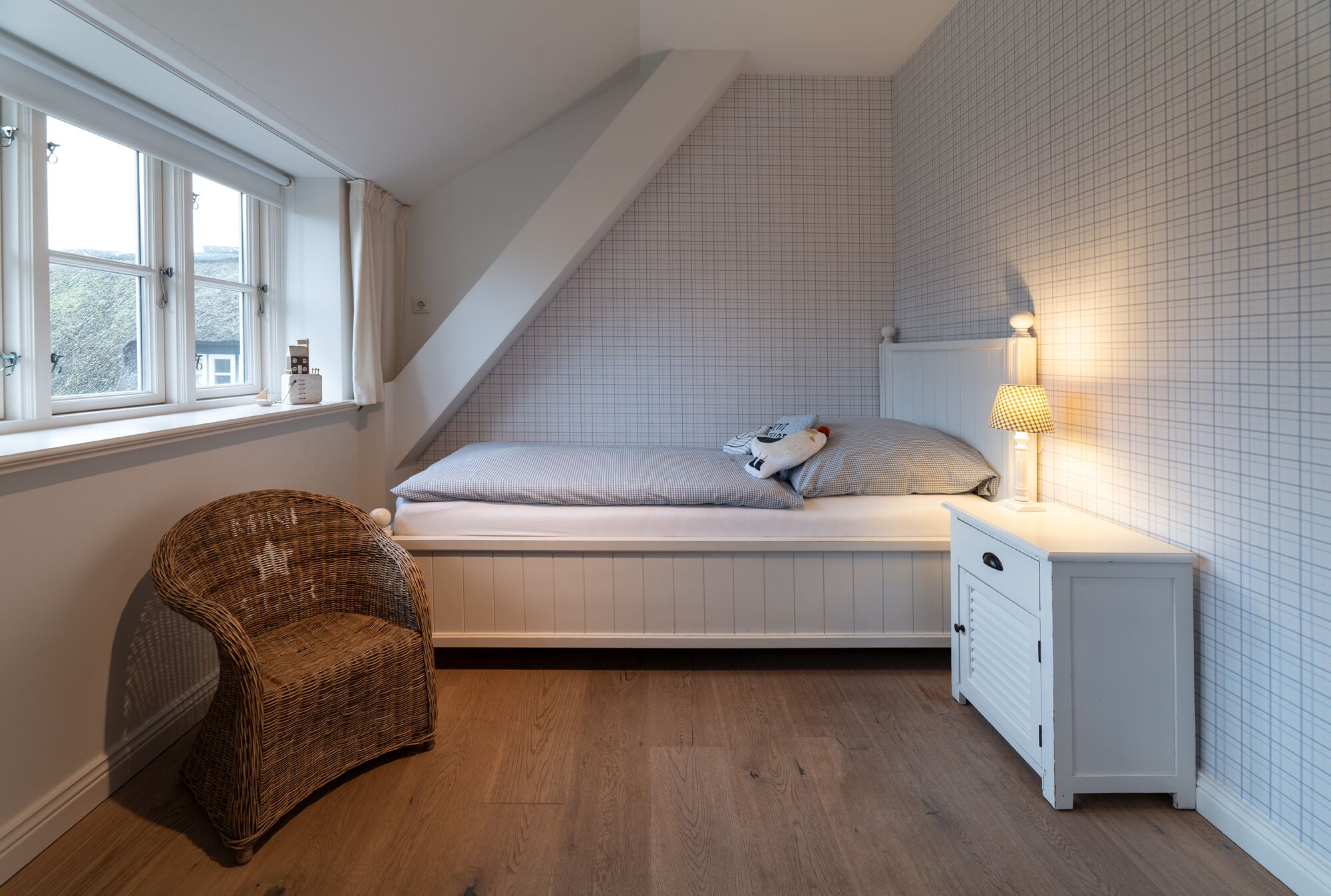 A bedroom with a sloped ceiling, a bed with pillows, a wicker chair, a nightstand with a lamp, and a window with curtains.
