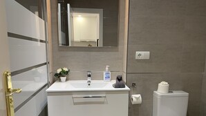 Et moderne badeværelse med en hvid vask, spejl og toilet. Der er to skuffer under vasken, en potteplante og en håndspritflaske.
