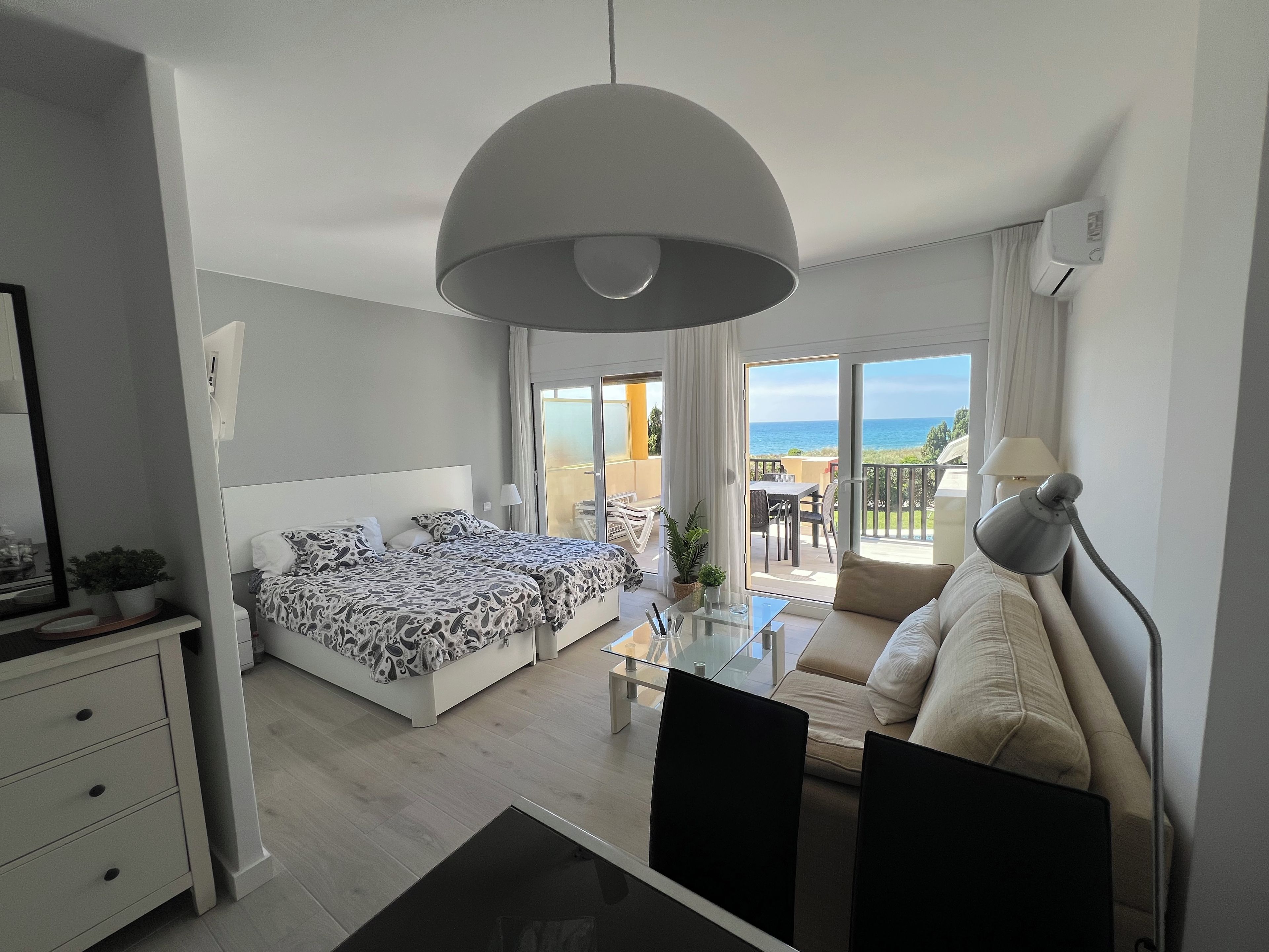 A modern hotel room with a bed, a sofa, a dining table, and a balcony with a view.