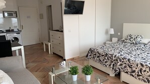 Et moderne hotelværelse med seng, sofa, sofabord i glas med planter, fjernsyn og et tekøkken med mikrobølgeovn og vaskemaskine.