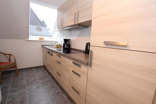 A kitchen with light wood cabinets, a black countertop, and a wicker chair. - Ferienwohnung in Westerland auf Sylt (Westerland)