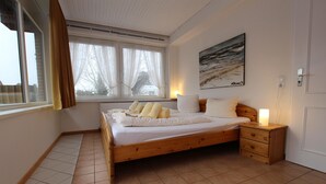 A bedroom with a wooden bed, a bedside table with a lamp, a window with curtains, and a painting on the wall. - Ferienwohnung in Westerland auf Sylt (Westerland)
