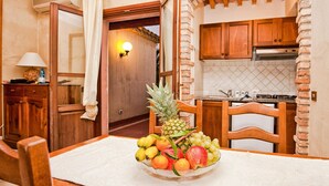 A dining area with a table set for two, a fruit bowl on the table, wooden chairs, and a view of a kitchen with brick walls and wooden cabinets. - 1 bedroom cozy apartment in Pula (CA) (Pula (CA))