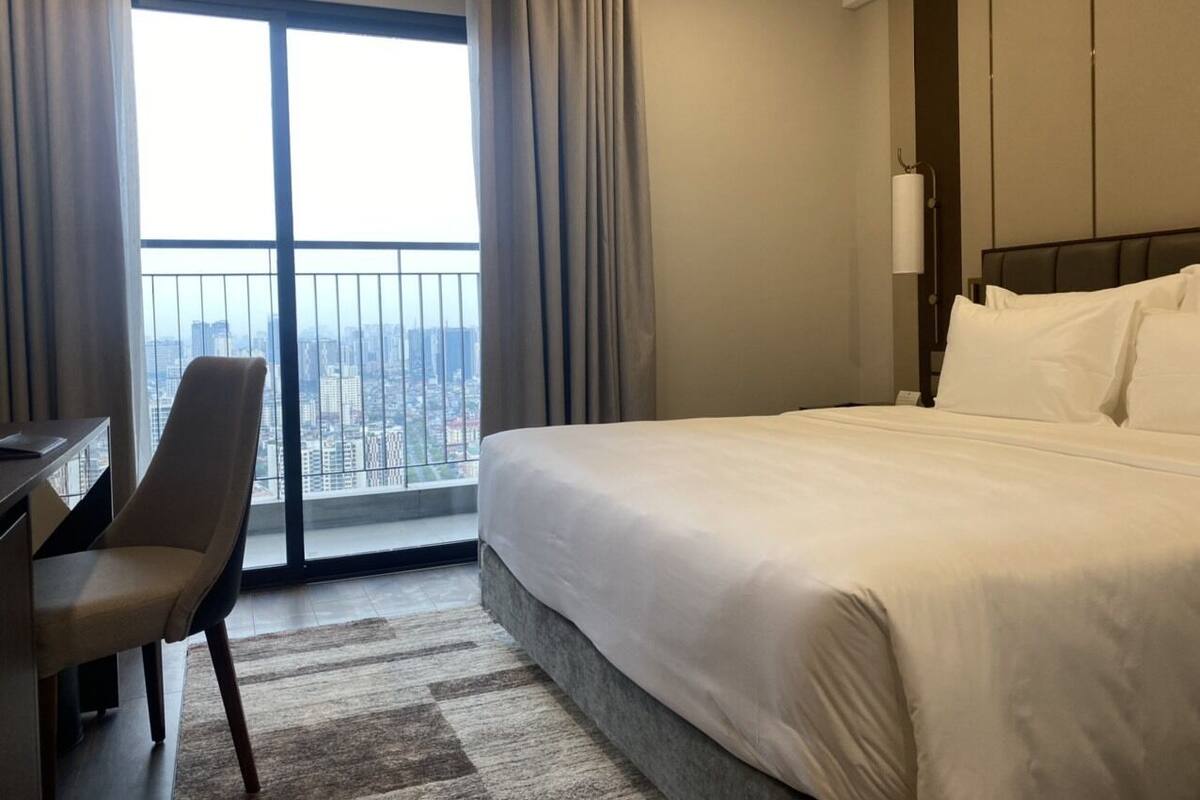 Une chambre d’hôtel avec un grand lit, un bureau et une chaise. La fenêtre offre une vue sur le paysage urbain.