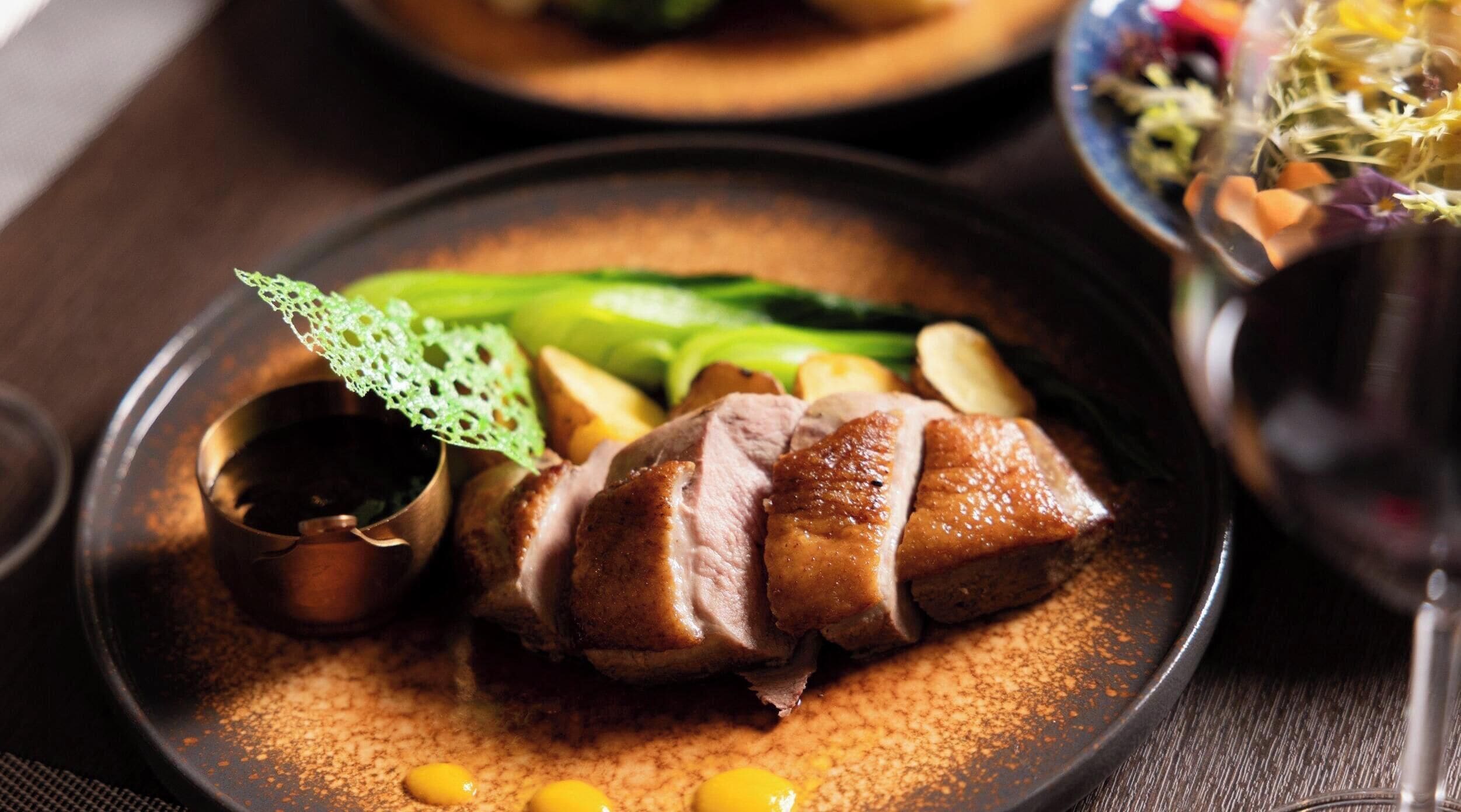 A plate of food with a duck, vegetables, and a small dish of sauce.