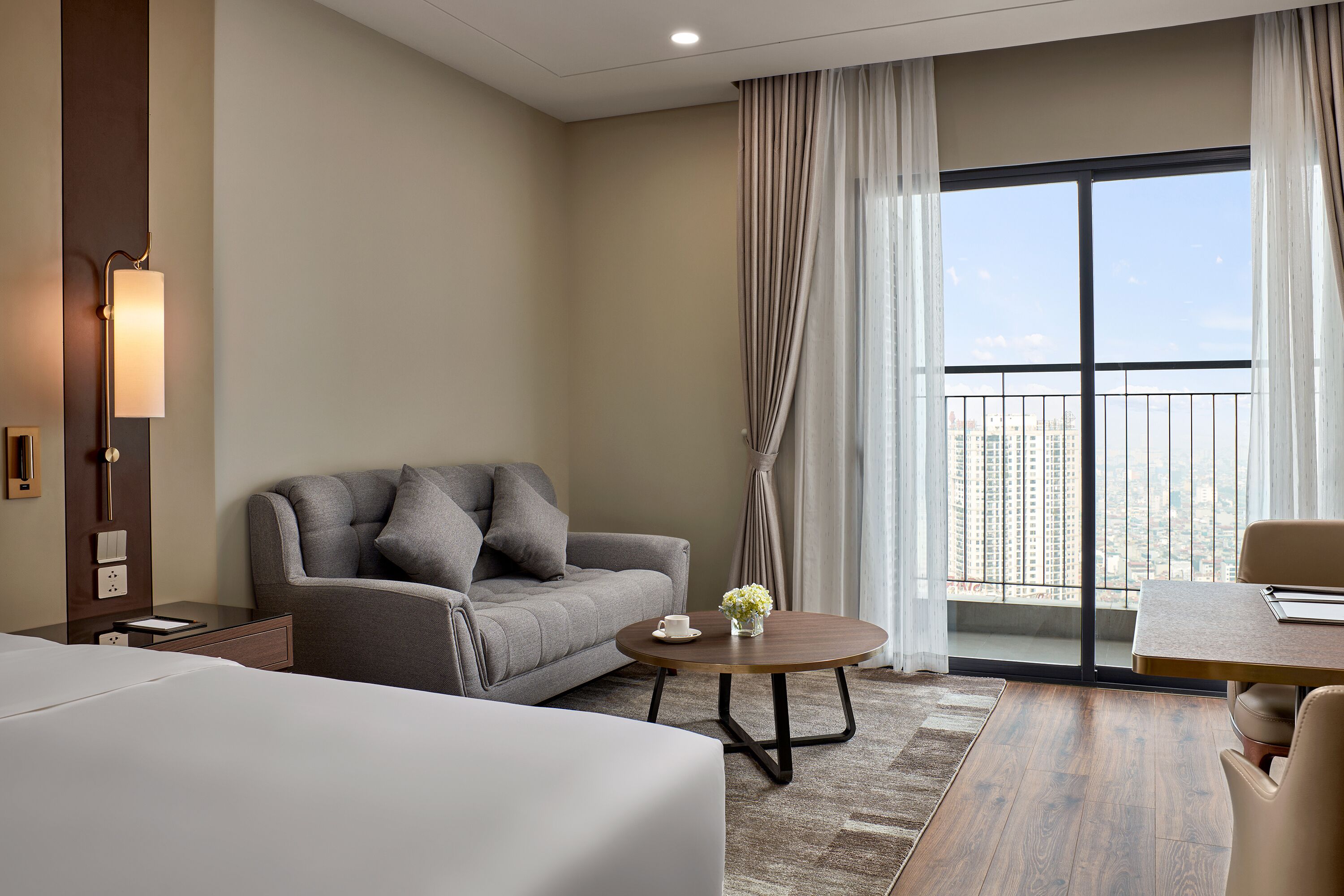 A modern hotel room with a large bed, a grey sofa, a round coffee table, and a view of the city through a large window.
