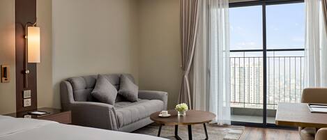 A modern hotel room with a large bed, a grey sofa, a round coffee table, and a view of the city through a large window.