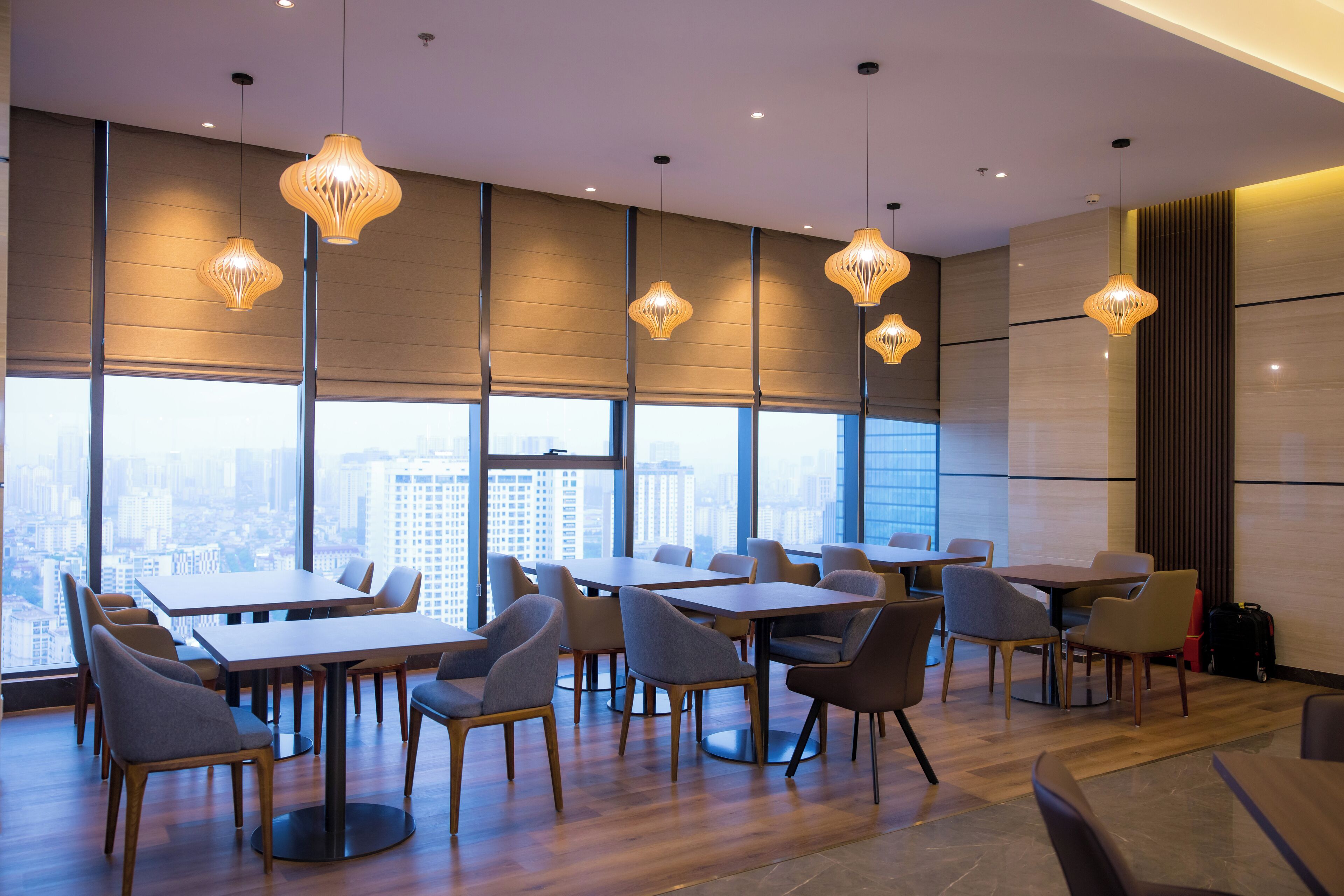 A modern hotel dining area with tables and chairs, large windows with a city view, and pendant lighting.