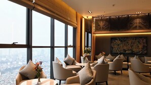 A high-rise hotel lobby with large windows offering city views, comfortable seating areas, and a decorative wall with a large abstract painting.