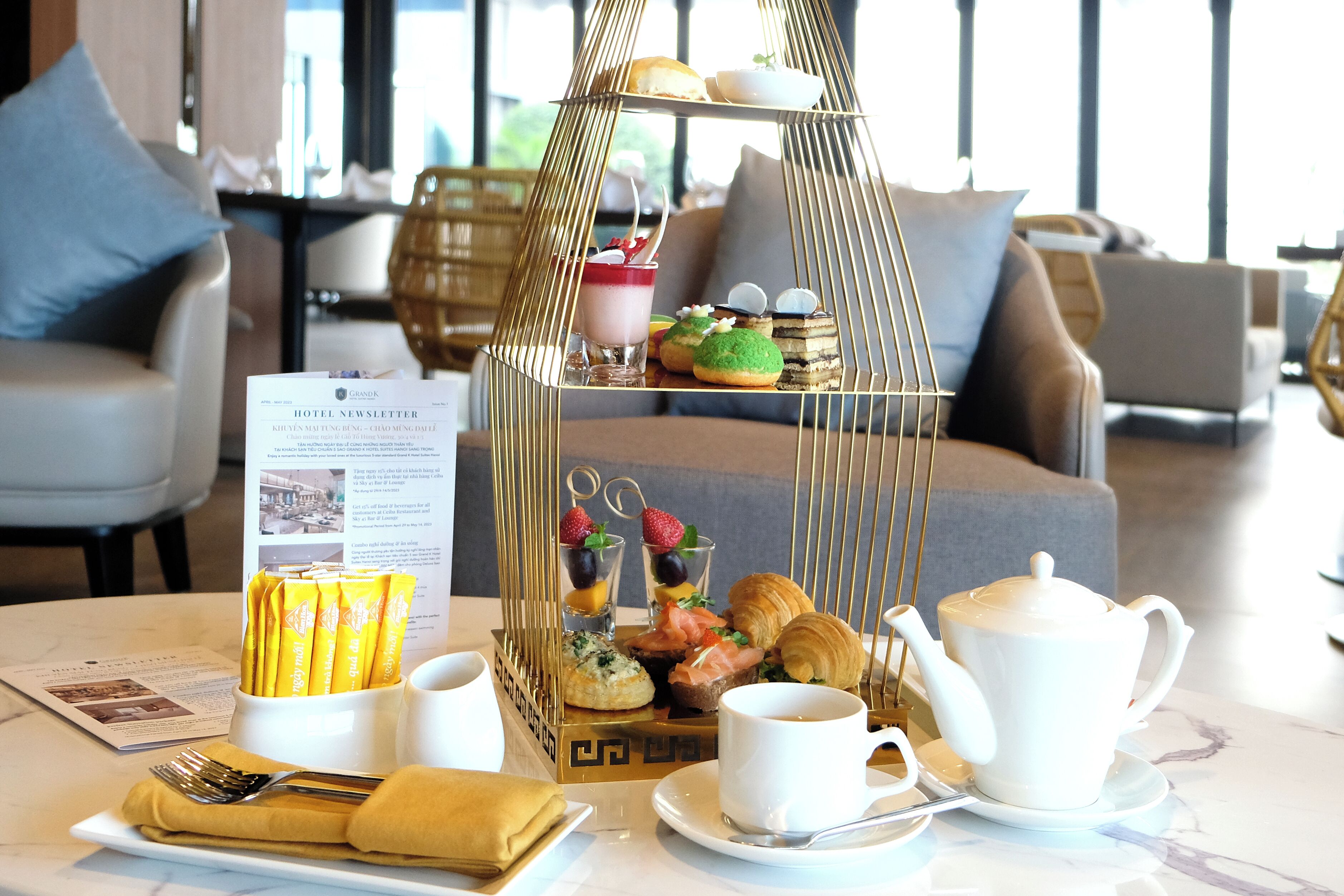 A breakfast setup with a tiered serving tray, a white teapot, cups, and a menu on a marble table in a modern room with grey sofas.