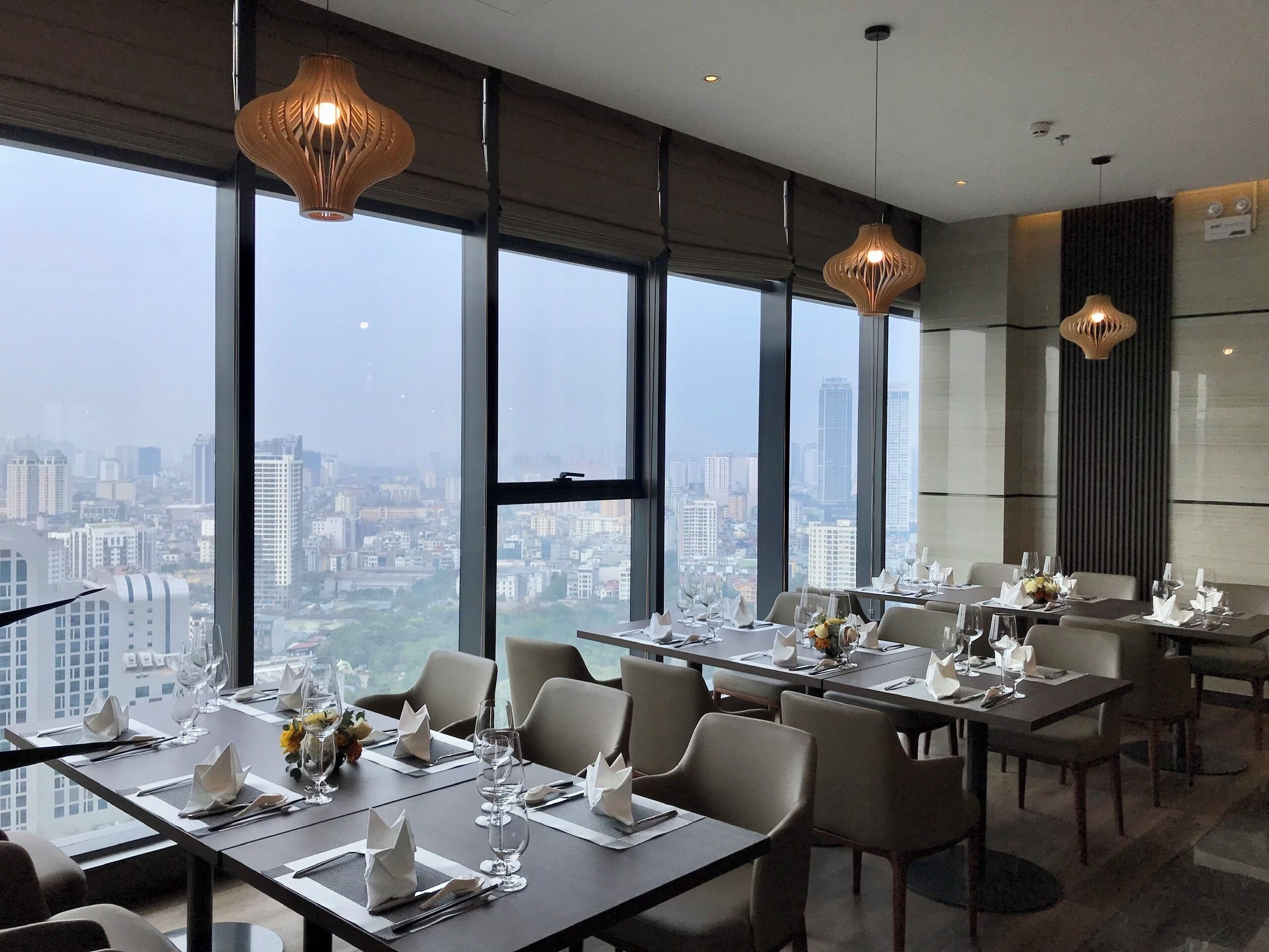 A modern dining area with tables set for service, large windows offering a city view, and pendant lighting.