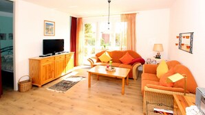 A living room with a sofa, a coffee table, a TV on a wooden cabinet, and a window with curtains. - Ferienhaus, Dornumersiel (Dornumersiel)