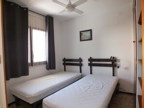 A room with two single beds, a ceiling fan, a window with curtains, and a painting on the wall.