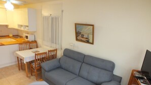 A living room with a grey sofa, a dining table with chairs, a kitchen area with cabinets, and a painting on the wall.