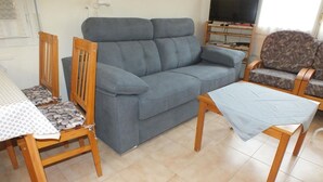 A living room with a grey sofa, wooden chairs, a small table, and a television on a stand.