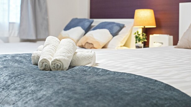 A neatly made bed with rolled white towels, a blue blanket, and a bedside lamp. - HOME Tengachaya2F (Osaka)