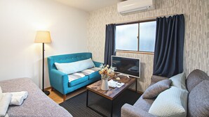 A hotel room with a blue sofa, a grey sofa, a wooden coffee table with a vase of flowers, a flat-screen TV, and an air conditioning unit. - HOME Tengachaya2F (Osaka)