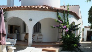 A courtyard with white lounge chairs, a small table, and a bench. There are steps leading to an entrance, and a plant with purple flowers is visible.