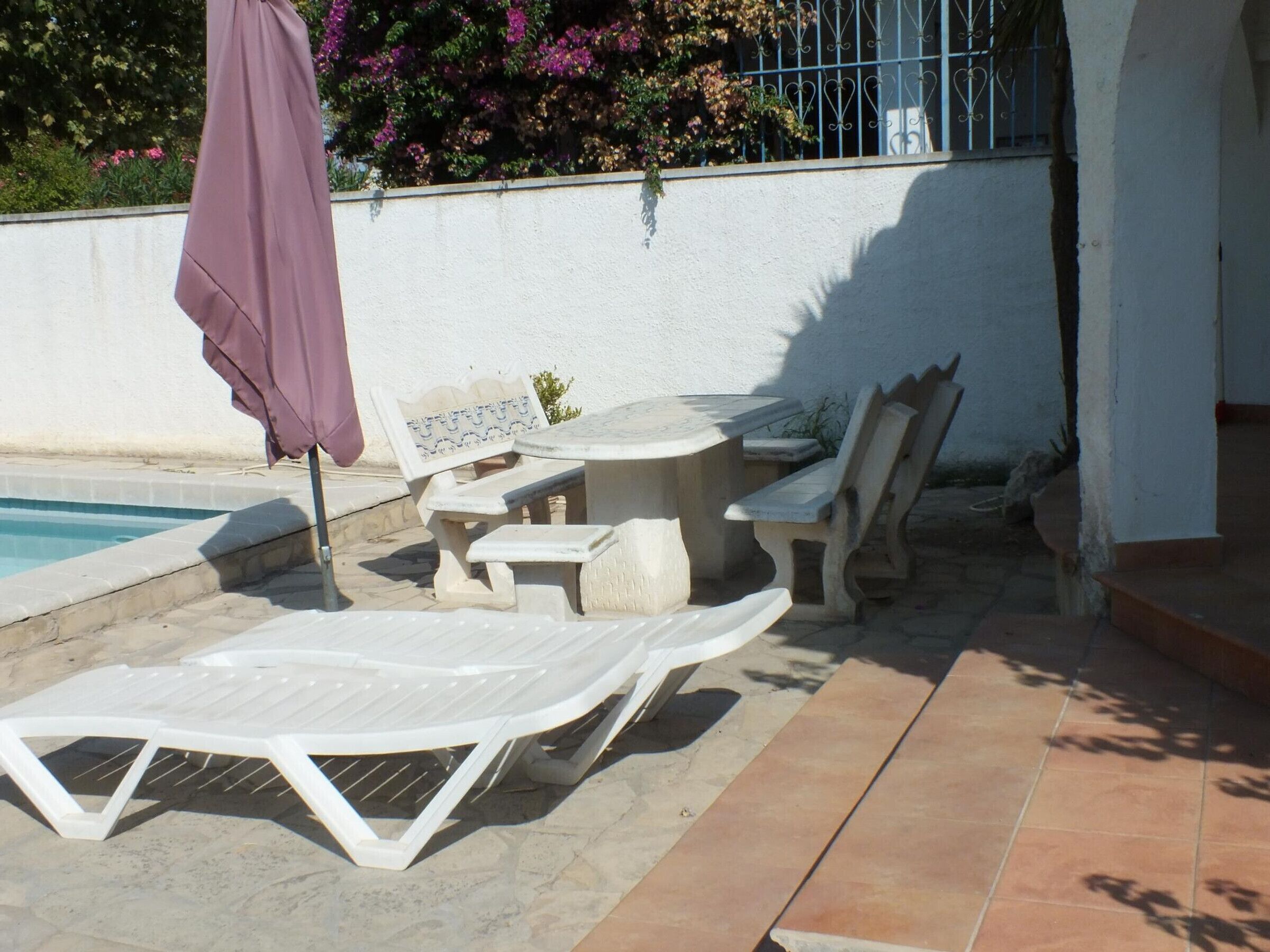 A poolside area with a table and chairs, a lounge chair, and a purple umbrella.