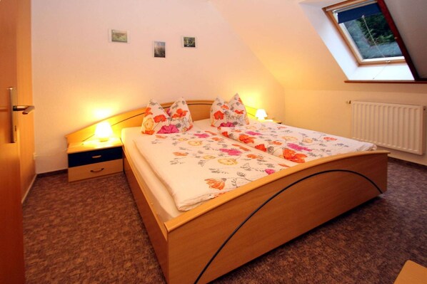Un dormitorio con cama de madera, ropa de cama floral, una mesita de noche con lámpara, un cajón pequeño y un tragaluz.