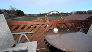 A wooden deck with a round table and two chairs, a small wreath on the railing, and a view of a grassy field and houses in the distance.