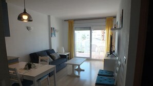 Una sala de estar con sofá, una mesa de comedor y televisor. Hay un balcón con cortinas amarillas y vistas al exterior.
