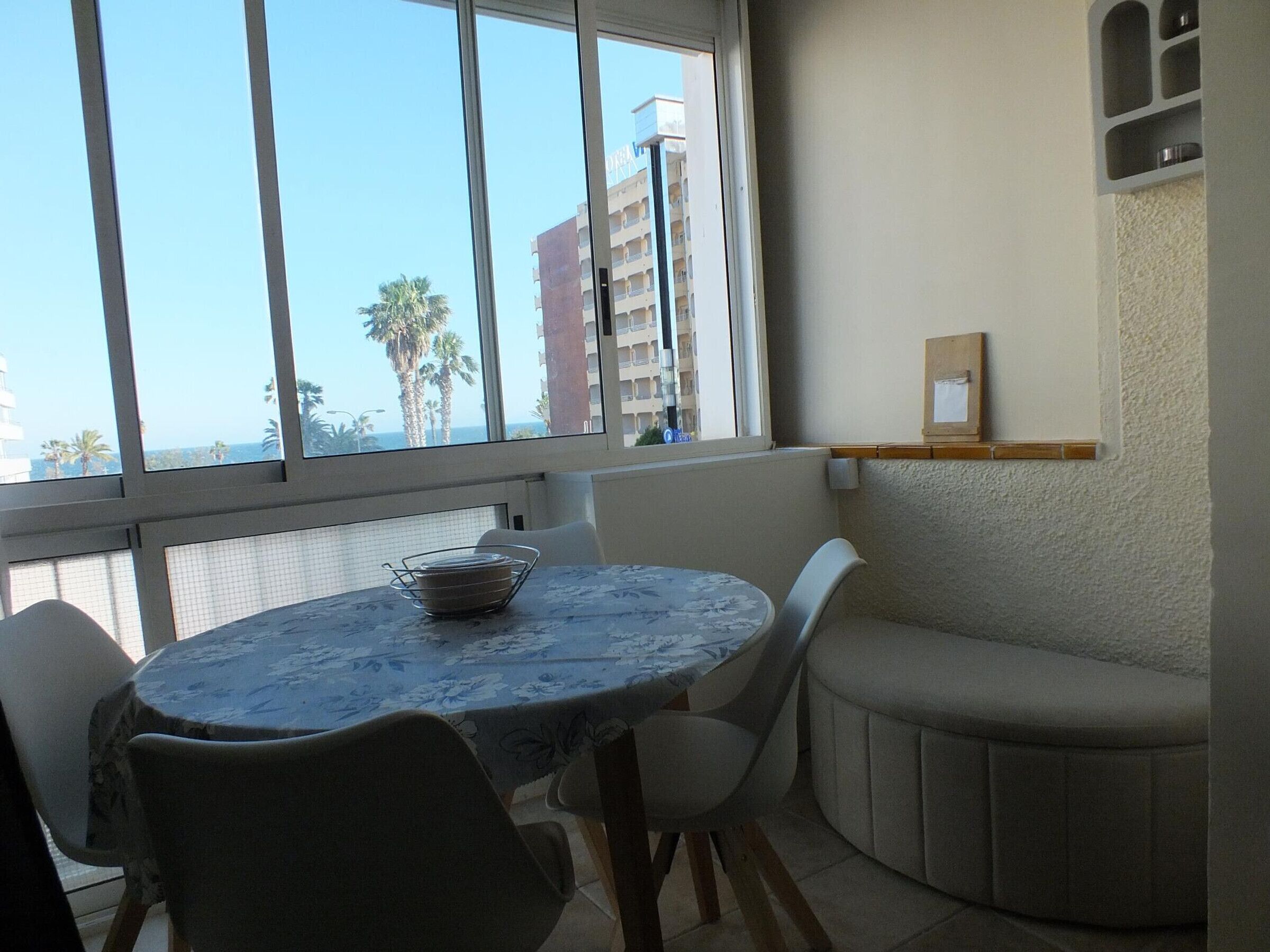 Zona pranzo con tavolo rotondo, sedie, una piccola panca e una finestra vista palme e edifici.