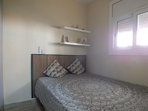 Une chambre Ă coucher avec une couvre-lit Ă motifs, une tĂȘte de lit Ă rayures verticales, deux oreillers, une table de chevet avec une lampe et une fenĂȘtre avec des stores.