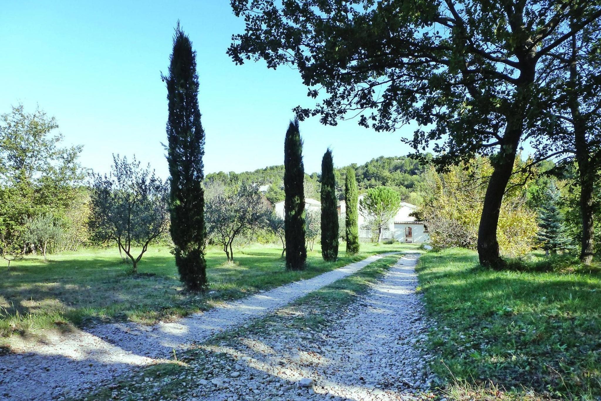 Ein Kiesweg führt durch einen Garten mit hohen Bäumen und einem Gebäude in der Ferne.