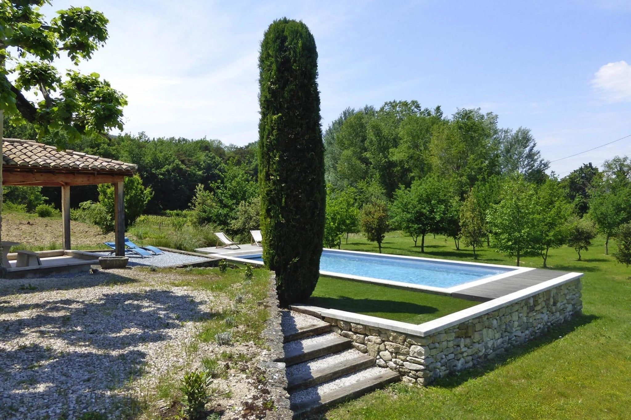 A swimming pool with a stone border, a tall conical hedge, and a pergola with a tiled roof.