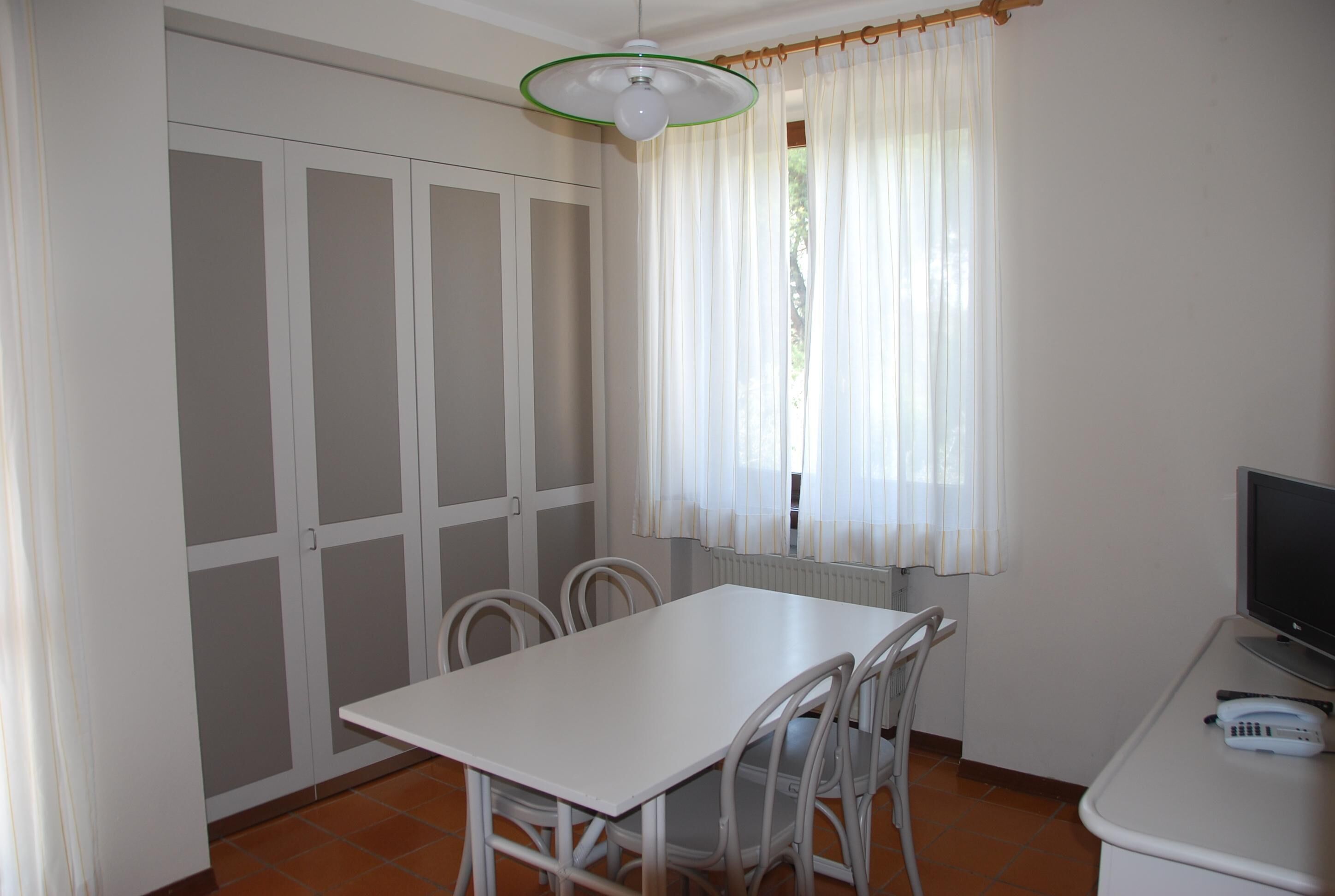 A room with a white table and chairs, a television, and a window with sheer curtains.