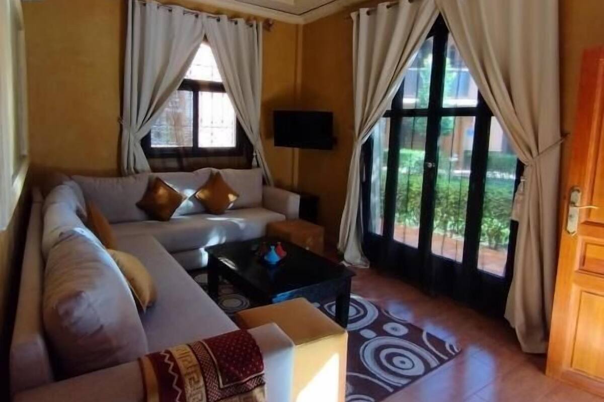 A living room with a sofa, a coffee table, and a TV. There are curtains and a ceiling fan.