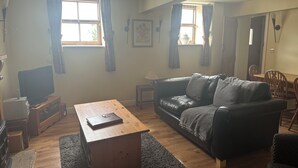 Una sala de estar con una mesa de centro de madera, un sofá de cuero negro, un televisor sobre un mueble de madera, y una zona de comedor con sillas y una mesa de madera.