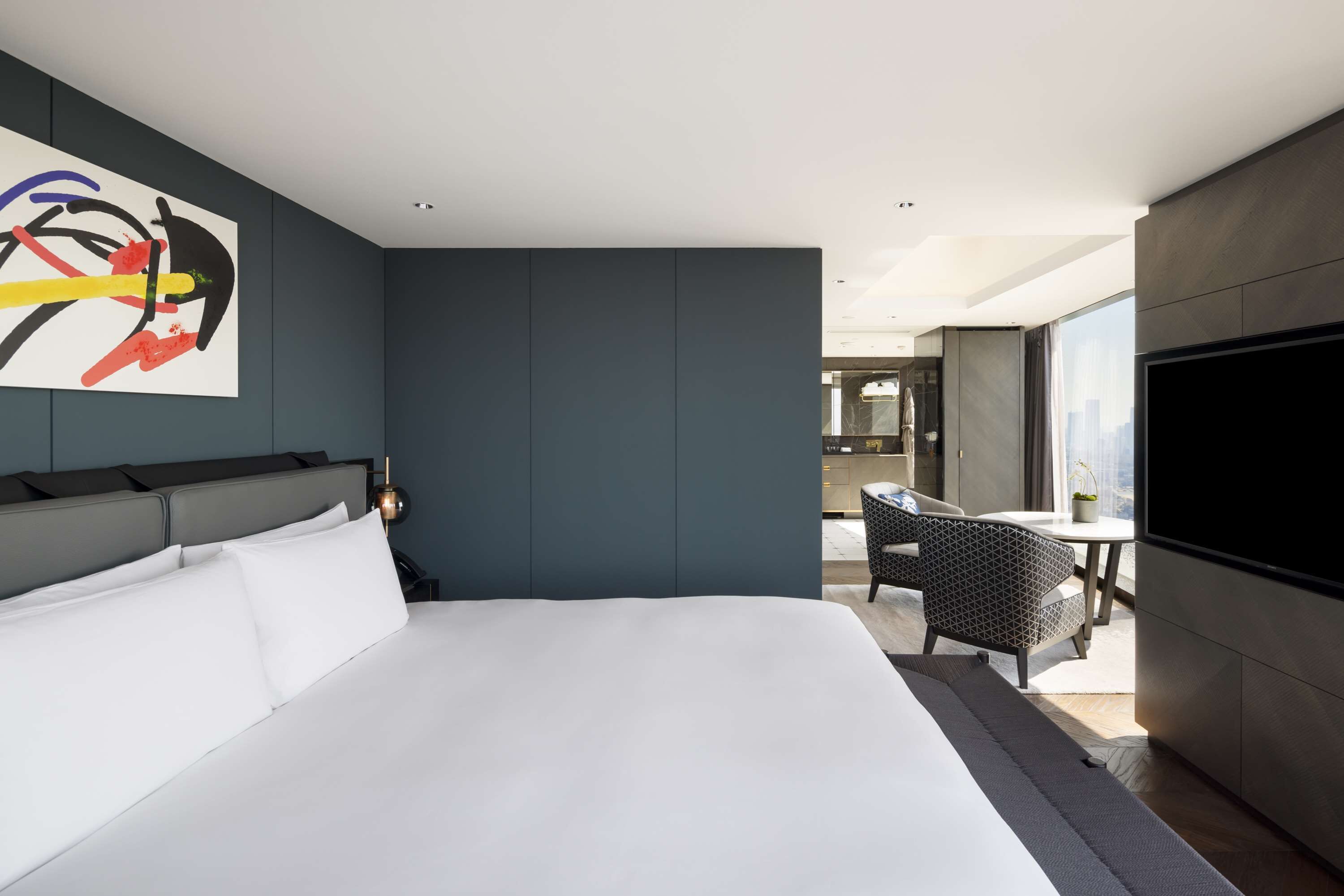 A modern hotel room with a large bed, a flat-screen TV, and a dining area with a table and chairs.
