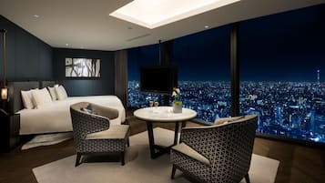 Una habitación de hotel moderna con una cama grande, dos sillones estampados, una mesa redonda y vistas a la ciudad a través de amplios ventanales.