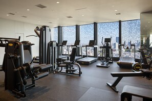 A modern gym with various exercise equipment, large windows offering a city view, and a spacious layout.