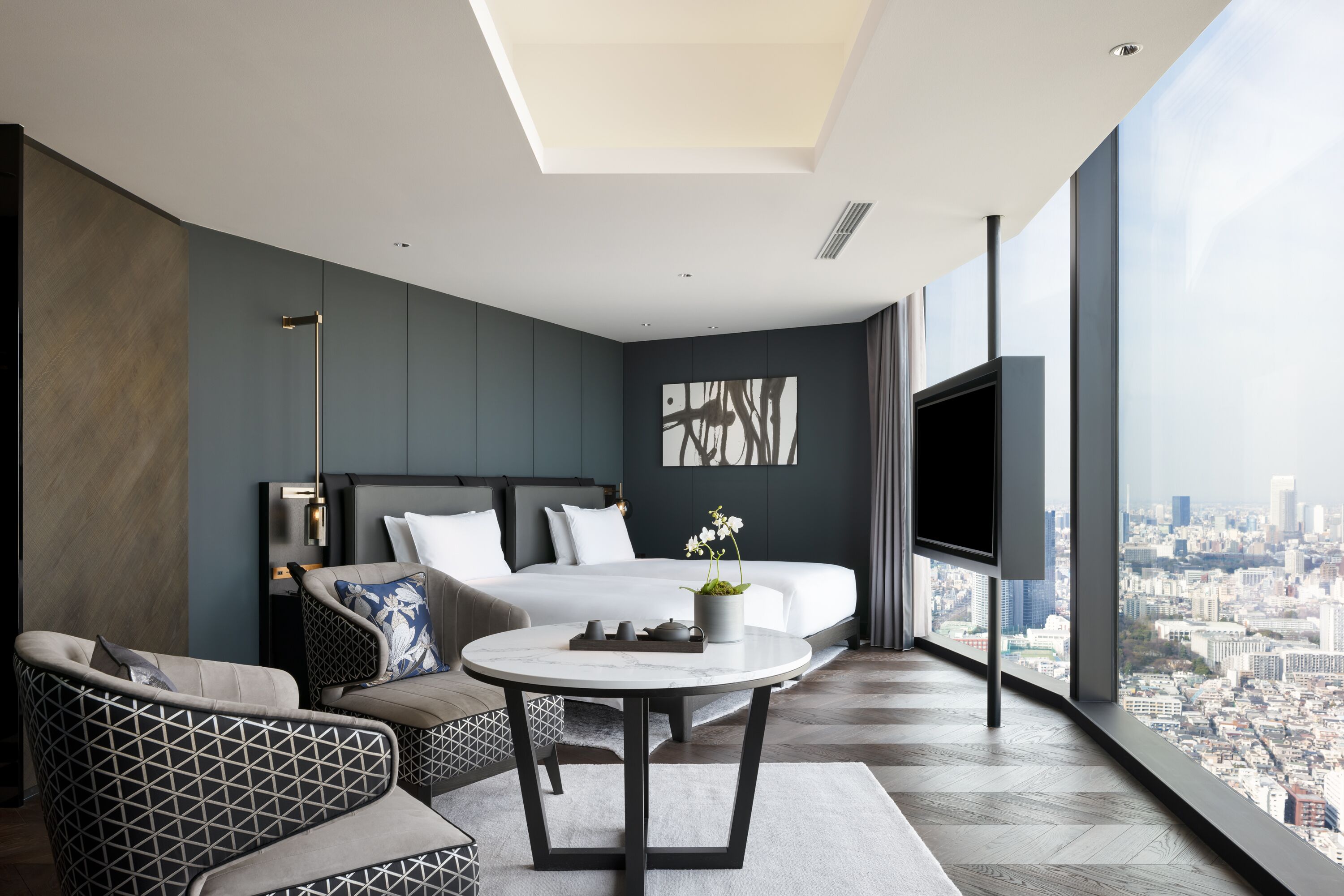 A modern hotel room with a large bed, a round table, a sofa, and a city view through large windows.